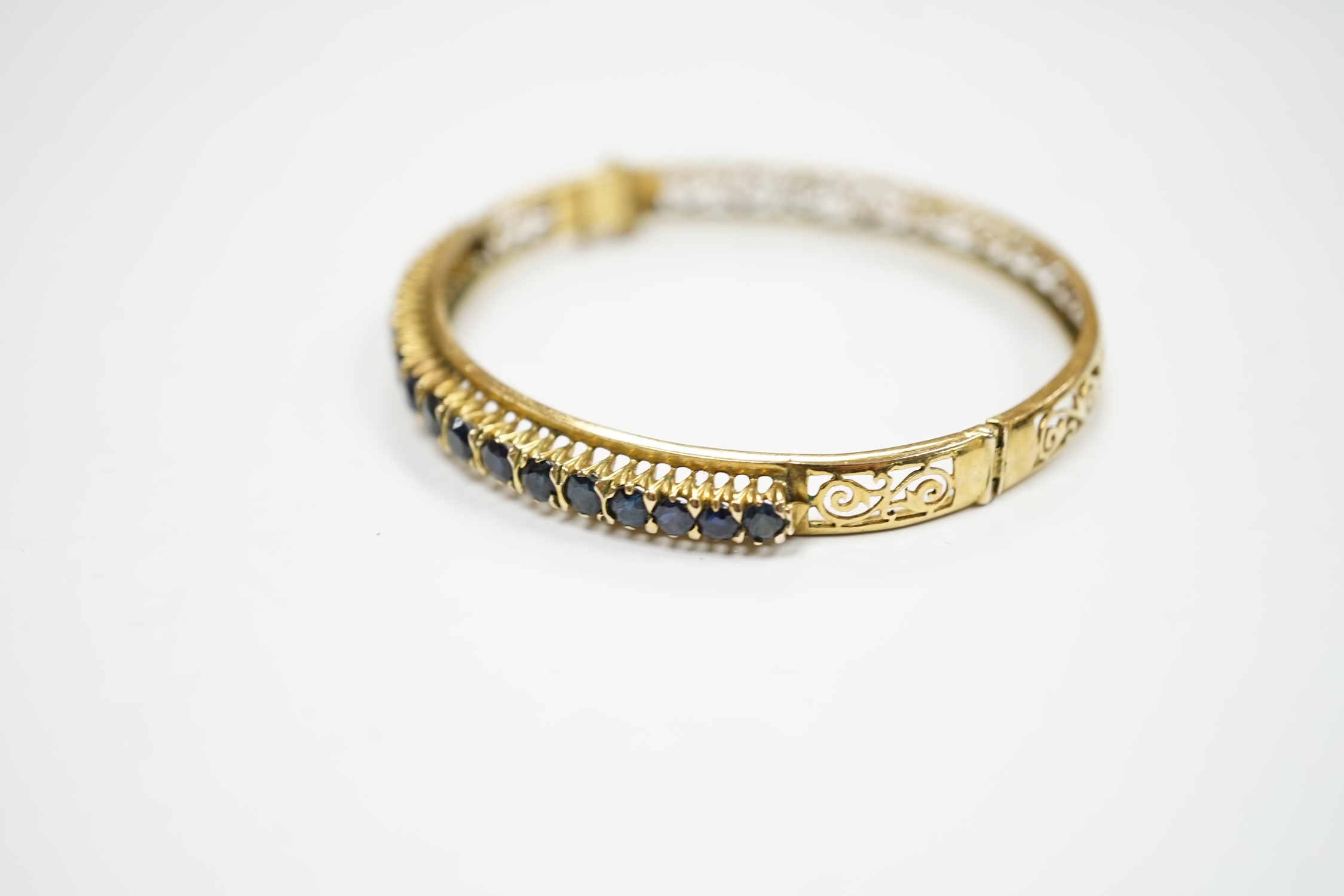 A pierced yellow metal and eighteen stone sapphire set hinged bangle, interior diameter 58mm, gross weight 14.1 grams.